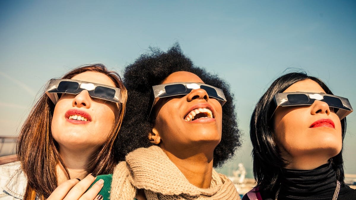 Three women wearing solar eclipse glasses looking up at the sky